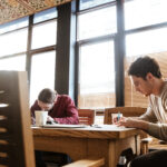 Photo of two attractive young colleagues in office working while writing notes at notebook. Coworking.